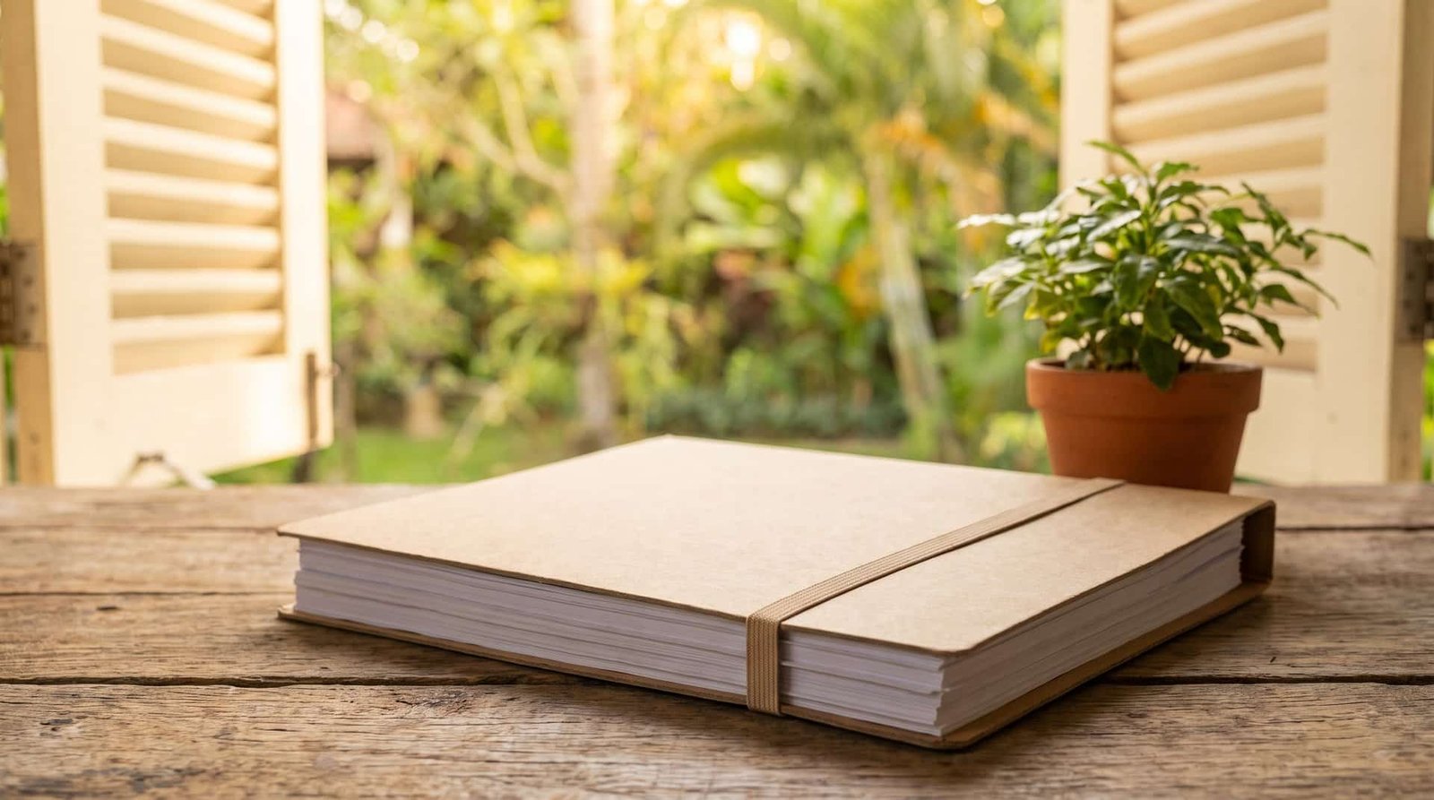 Dossier cartonné kraft fermé par un élastique, posé sur une table en bois devant une fenêtre ouverte sur un jardin tropical